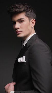 sexy young businessman in black tuxedo moving, posing from behind while adjusting tux and looking to side in front of dark grey background with flash lights on