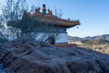 A guard tower at an ancient palace complex in China as seen from the shady area between stacked of raw hard rocks and trees under clear blue sky.