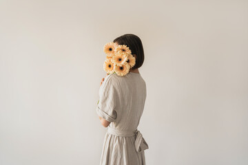 Beautiful young woman in neutral sandy linen dress holding gerber flowers bouquet over white wall....