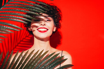 Fashion beauty portrait of young smiling brunette woman with evening stylish  makeup. Sexy model with curly short hair posing in studio. With red bright lips. Tropical palm leaf. In sunglasses