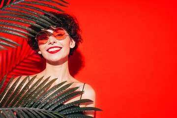 Fashion beauty portrait of young smiling brunette woman with evening stylish  makeup. Sexy model with curly short hair posing in studio. With red bright lips. Tropical palm leaf. In sunglasses