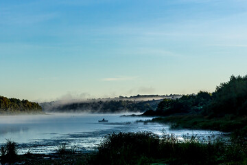 Fishermen on  boats early in the morning at dawn in the fog or mist at golden hour catch fish on the Oka River, Russia. Green grass and trees, a river bend against a clear light sky in summer.