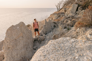 Man in a T-shirt and shorts with backpack staying on rocky coastline with warm sun light.
