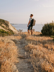 Fototapeta premium Man in a T-shirt and shorts with backpack on the coastline with warm sunset.
