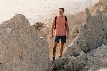 Man in shorts with backpack staying on rocky coastline with warm sun light.