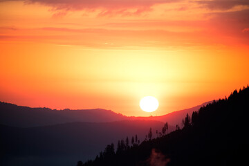 Silhouettes of mountain ranges and trees against the backdrop of the setting sun.
