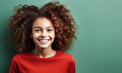 Fototapeta premium Delightful young girl with a captivating smile and playful curly hair in a bright red shirt against a soft mint green background