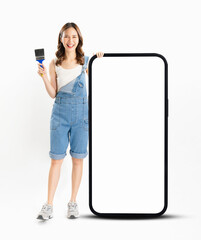 Appealing Asian woman holds paintbrushes and a big smartphone with a blank screen against a white...
