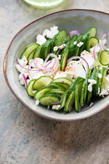 Salad with fresh cucumber, feta cheese and red onion in a grey bowl, vertical shot on a light-brown granite background, middle close-up