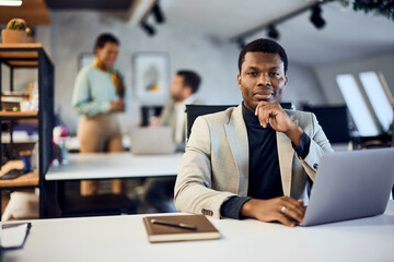 Portrait of a black businessman, working at the office, using a laptop, and looking at the camera.