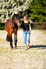 Young woman with a black shirt and short highlighted hair stands with her horse on a sunny riding arena.