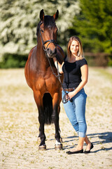 Obraz premium Young woman with a black shirt and short highlighted hair stands with her horse on a sunny riding arena.