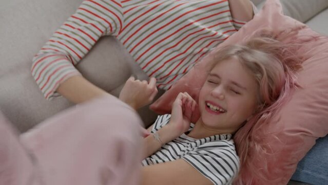An unrecognizable young woman playing with a laughing rose-haired girl lying on her knees while sitting on a light sofa.