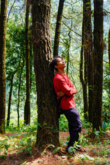 Javanese man in red is leaning against a tree in the forest. 