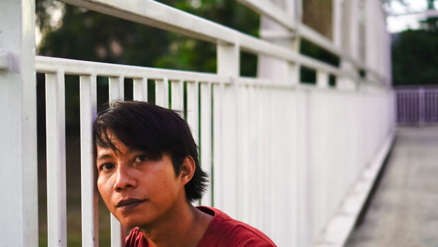 Portrait of Asian man in red shirt standing on white fence with pensive expression.