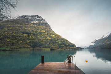 A small, empty wooden pier on the green waters of a lake in the Norwegian fjords, with snow-capped...