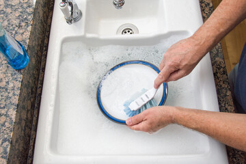 Washing dishes. A mans hands are visible as he washing a plate in a white sink with lots of...