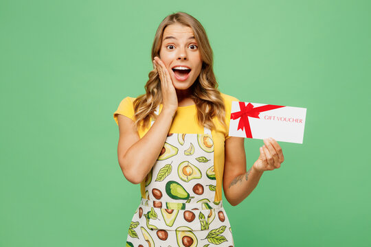 Young housewife housekeeper chef cook baker woman wear apron yellow t-shirt hold gift certificate coupon voucher card for store isolated on plain pastel green background studio. Cooking food concept.