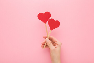 Young adult woman hand holding two red paper heart shapes on wooden sticks on light pink table background. Pastel color. Love concept. Closeup. Top down view. - obrazy, fototapety, plakaty