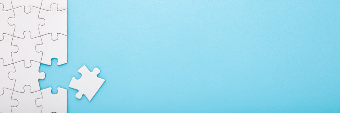 Unfinished white puzzle with last one piece on light blue table background. Pastel color. Wide banner. Closeup. Empty place for text. Top down view.