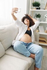 Pregnant woman blogger sits on the couch at home and takes pictures of herself on the phone, selfie and video call, consultation with the doctor online, pregnancy management