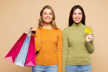 Young happy friend two women they wear orange green shirt casual clothes hold in hand credit bank card shopping paper package bags isolated on plain beige background Black Friday sale buy day concept - obrazy, fototapety, plakaty