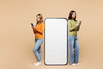 Full body side view young friends two women they wear orange green shirt casual clothes together big huge blank screen mobile cell phone with area using smartphone isolated on plain beige background.