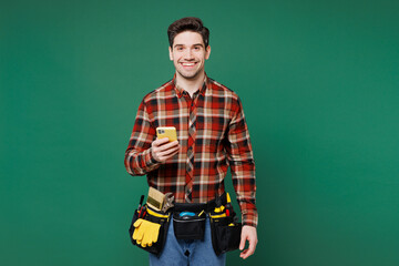 Young employee laborer handyman man wears red shirt hold in hand use mobile cell phone isolated on plain green background. Instruments accessories for renovation apartment room. Repair home concept.