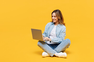 Fototapeta na wymiar Full body smiling happy young IT woman wear blue shirt white t-shirt casual clothes sitting hold use work on laptop pc computer isolated on plain yellow background studio portrait. Lifestyle concept.