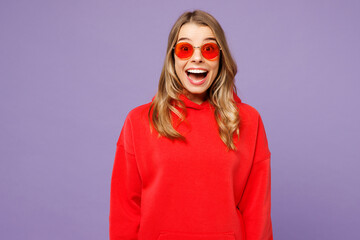 Young surprised excited overjoyed shocked blonde woman she wearing red hoody casual clothes sunglasses look camera isolated on plain pastel light purple background studio portrait. Lifestyle concept.