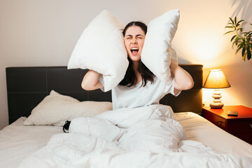 Portrait of annoyance Caucasian woman covers her ears with pillows and screams sitting in bed....