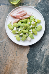 Potato gnocchi served with green pea sauce and bacon on a white plate, flat lay on a beige and grey granite background, vertical shot, copy space
