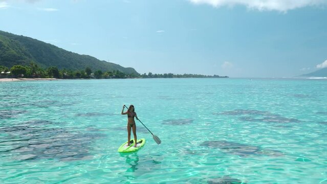 Girl paddle boarding crystal sea water aerial view. Coral reef tropical island, blue sky, sunny day. Young female rows standing on green paddleboard. Outdoor lifestyle travel, summer holiday vacation