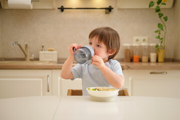 A child drinks from a non-watering cup while sitting at a table in a home kitchen. Cute little boy drinking from a bottle in the kitchen at home. Toddler baby two years old at dinner