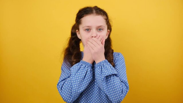 Portrait of shocked little caucasian girl child in blue dress cover palms mouth oops gesture, posing isolated over yellow color background wall in studio. Childhood lifestyle and emotion concept