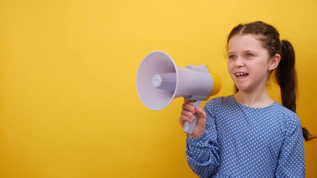 Portrait of excited little girl child 8-9 years old wearing blue dress screaming aside shouting hot news in megaphone, posing isolated on yellow background wall in studio. Childhood lifestyle concept