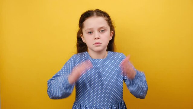 Portrait of confused little girl child 8-9 years old in blue dress show spread hand oops gesture, posing isolated over light yellow color background wall in studio. Childhood lifestyle concept