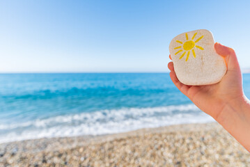 Child's hand holding a beach stone with a sun drawn on it, a defocused beach in the background.