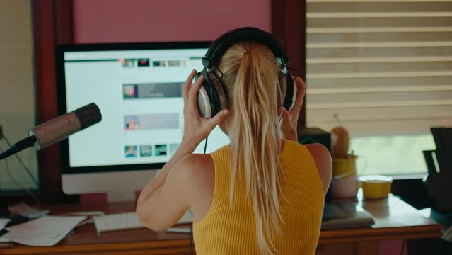Female content creator or podcaster take headphones on and work on her audio project at home studio. Freelance graphic designer or illustration artist at computer workstation. 