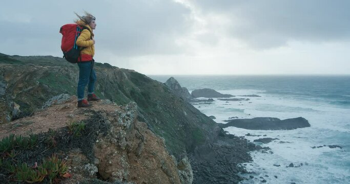 Cinematic shot of young female hiker stands on the top of the cliff alongside ocean. Young woman turns and looks around. Inspiring travel explorer reaching new horizons. Outdoor lifestyle. 