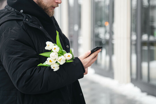 A man is using a smartphone and holding a bouquet of white tulips outside. Flower delivery concept, Women's day, Valentine's day, copy space.