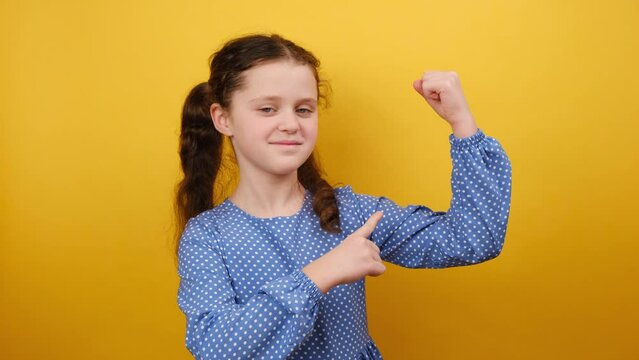 Portrait of strong sporty little girl kid 8-9 years old wearing blue dress point index finger on biceps muscles on hand, posing isolated over yellow color background wall in studio. Childhood concept