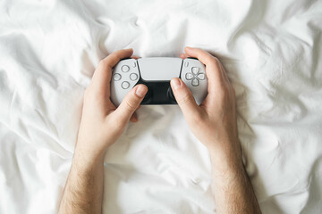 Man playing video games with game controller while lying in bed on white sheets, top view, gaming console concept.
