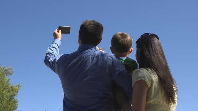 Young family with child taking photos with mobile phone and waving hands to the airplane flying over in the blue sky