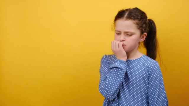 Portrait of pensive thoughtful little girl child 8-9 years old wearing blue dress look aside biting nails fingers, posing isolated over yellow background wall in studio. Childhood lifestyle concept