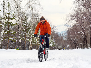 Fototapeta na wymiar A guy rides a bicycle in a winter park. A cyclist in an orange jacket rides with difficulty along a snowy path. Eco-transport in the city
