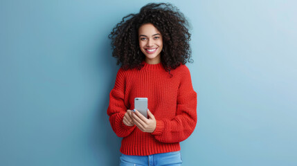 young woman with voluminous curly hair is smiling and holding a smartphone, wearing a red sweater, against a teal background