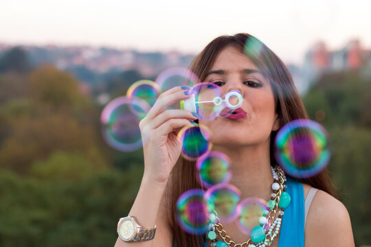 Young attractive brunette with necklace making bubbles outdoors in the park. Back to the childhood