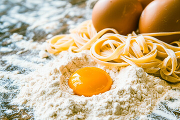 Close up shot of a mountain of flour with the yolk of an egg, fresh pasta and two eggs.