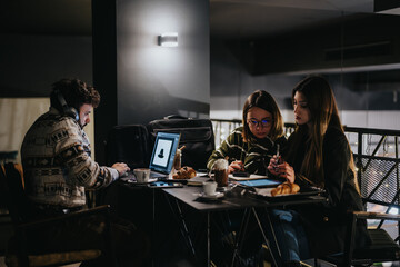 A group of three friends is engrossed in their work at night in a cafe, using laptops and taking...